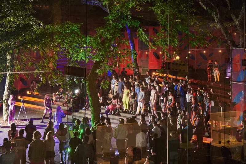 Large crowd gathers outdoors at night, illuminated by green and blue tree lighting. Performers on steps, red backdrop with geometric shapes, and string lights overhead.