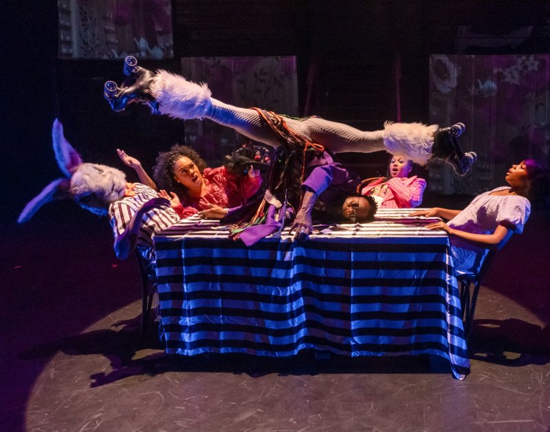 Performance art scene with figures around a striped table; one figure upside down wearing roller skates.