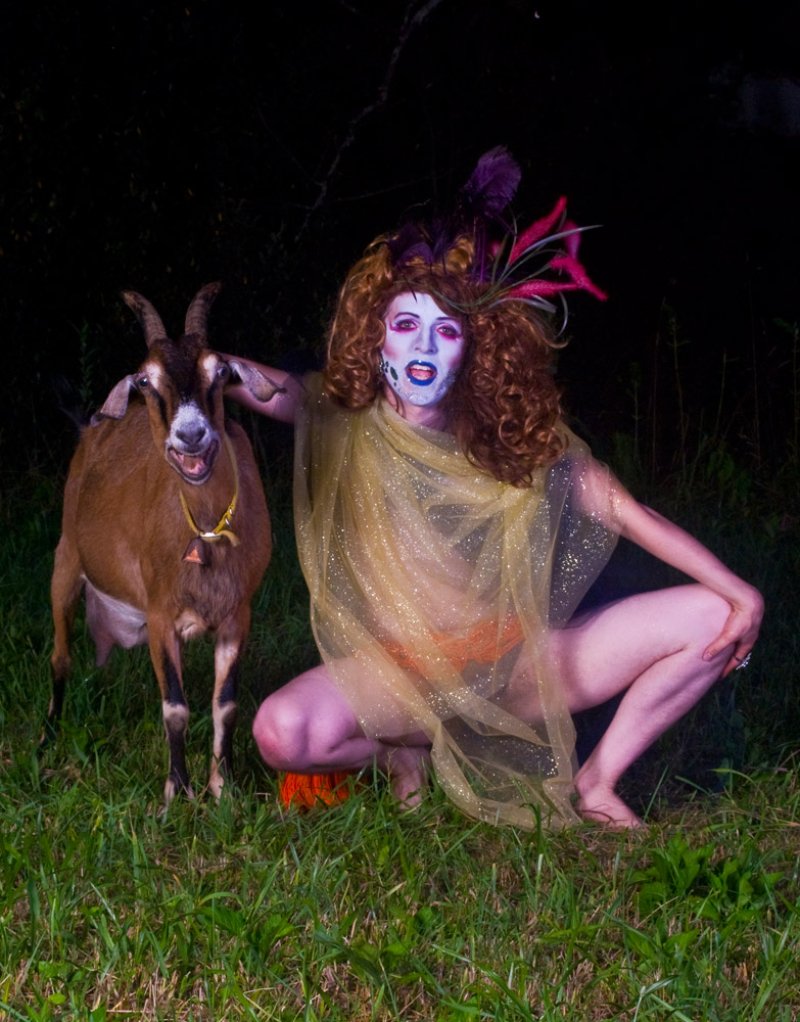 Drag performer with red hair, blue face paint, and sheer gold fabric squats next to a brown goat in dark grass.