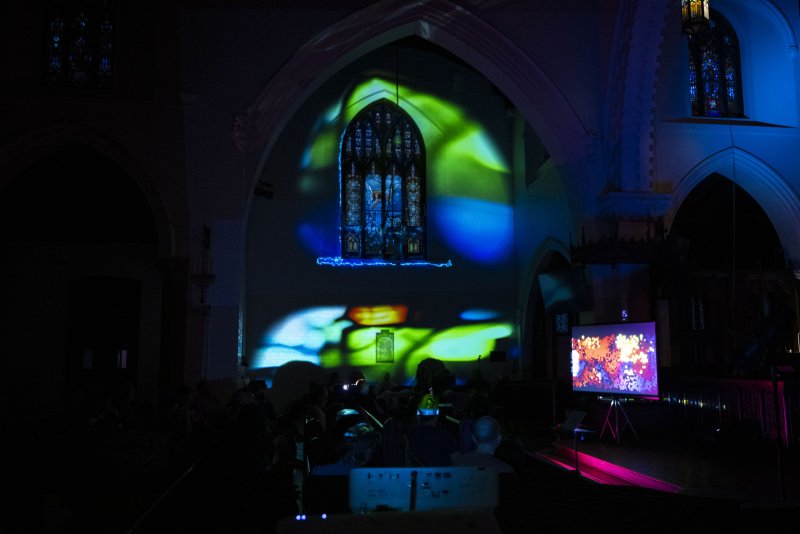 Dark church interior with green and blue light projections on an arched wall, a stained-glass window, and a screen displaying colorful abstract art.