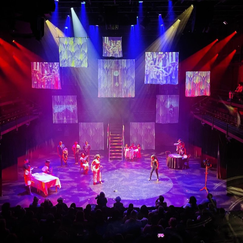 Theater stage with performers under red, white, and blue spotlights. Patterned screens hang above. Audience visible in the foreground.