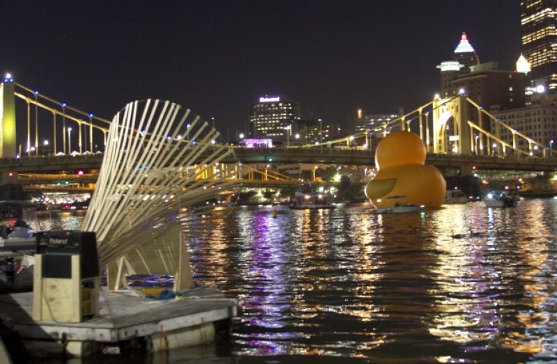 Night view of a river with a large yellow rubber duck and a wooden spiral sculpture, city lights reflecting on the water.