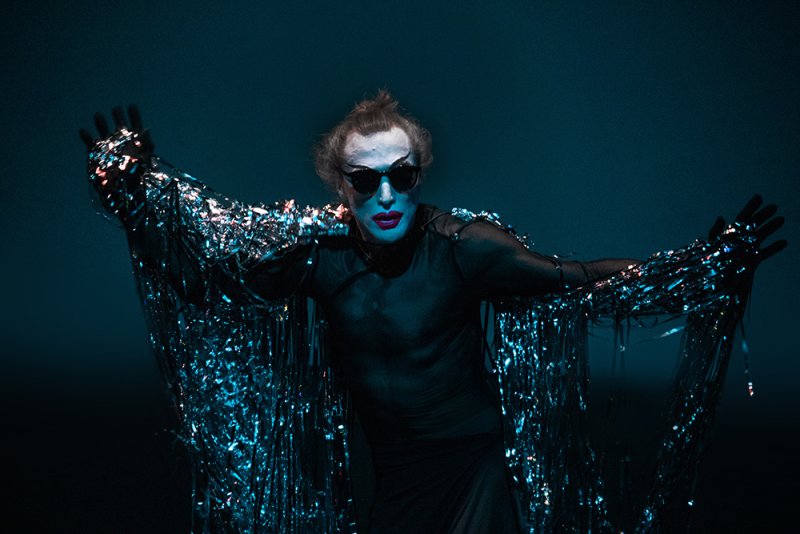 Performer with pale face, red lips, and sunglasses in black clothing. Silver tinsel drapes from their arms against a dark blue background.