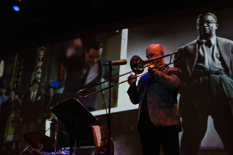 Bald man in a patterned suit jacket playing a trombone on a stage, with black and white projections of men in suits behind him.