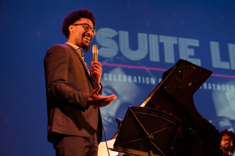 A man in a suit with glasses and curly hair smiles, holding a microphone, next to a grand piano on a stage with a blue background.