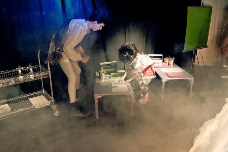 Two performers on a smoky stage, one standing in a white tunic, the other kneeling at a table with art supplies.
