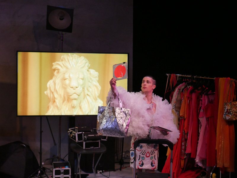 Performance shot featuring a figure in a large white ruffled garment, holding a red disc and a geometric bag. A screen displays a golden lion statue. Clothing rack with colorful garments visible in the background.