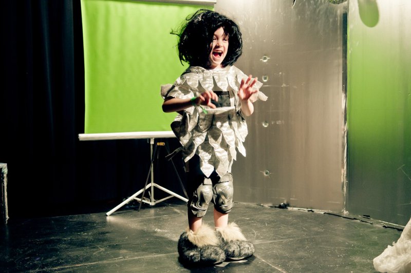 Child in a shiny, textured costume, black wig, and furry boots, standing on a dark stage with a green screen backdrop.