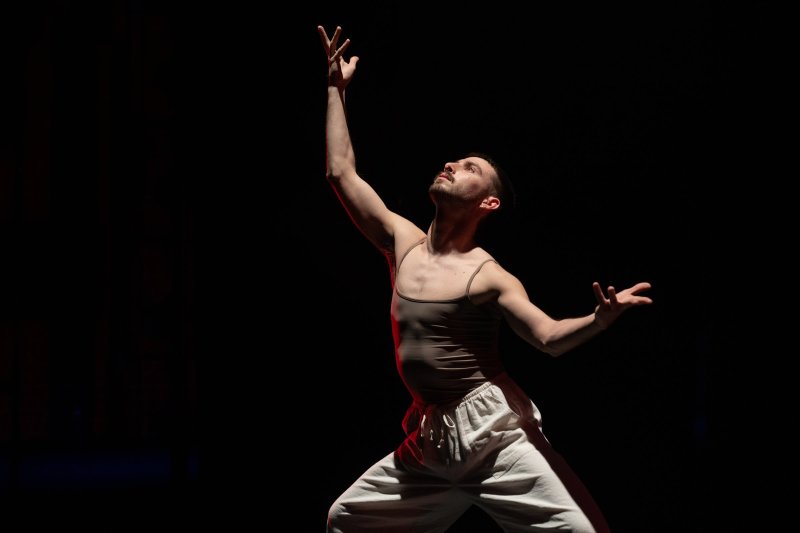 Dancer in a beige tank top and white pants, reaching upwards with one arm, bathed in warm light against a dark background.