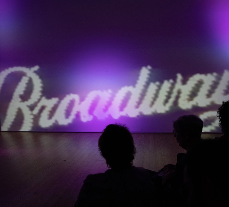 Broadway" projected in white, textured font on a purple wall, with silhouettes of people watching from a wooden floor.