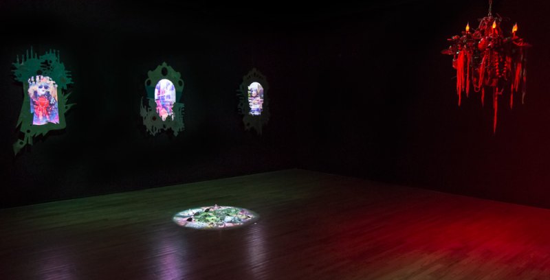 Dark gallery space with three wall-mounted screens displaying colorful, distorted figures. A red chandelier hangs to the right, and a circular projection illuminates the floor.