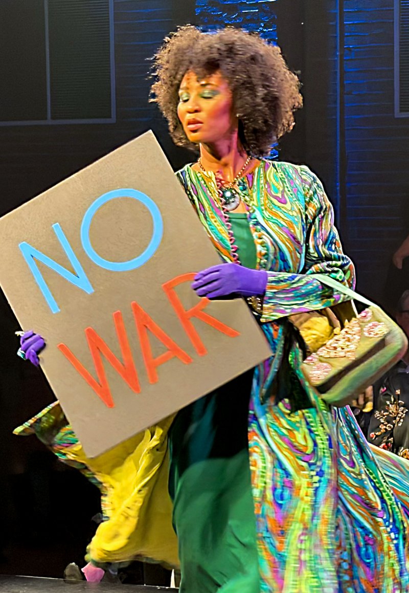 A woman with an afro holds a "NO WAR" sign, wearing a vibrant green, blue, and orange patterned dress with purple gloves.