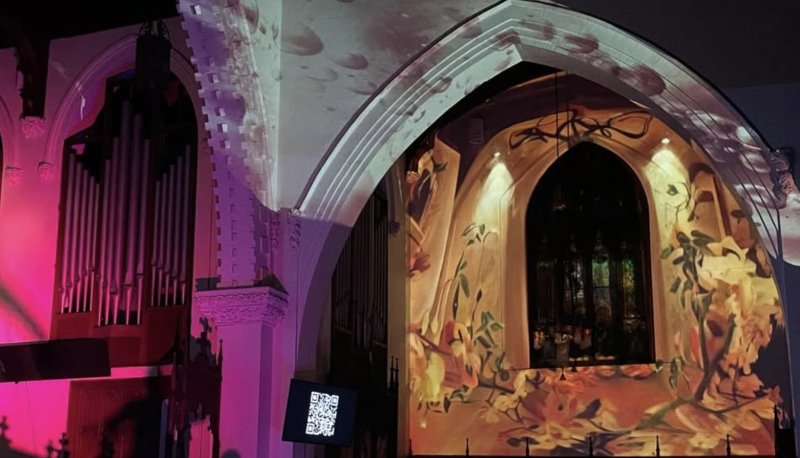 Digital projection of swirling autumn leaves on a church wall with a pipe organ and stained glass window, bathed in pink and purple light.