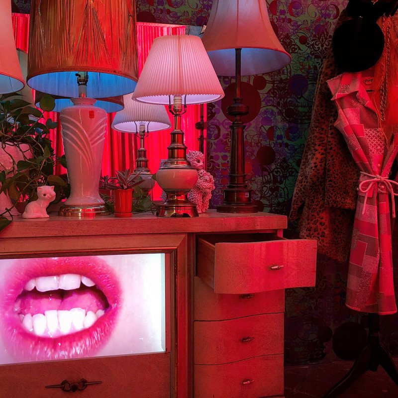 A dresser with lamps, plants, and a white cat figurine, bathed in red light, with a screen displaying an open mouth.