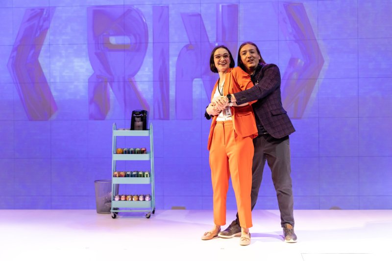 Two figures embrace on a stage with a blue backdrop featuring large, stylized letters. A woman in an orange suit stands in front, hugged by a man in a plaid blazer and grey pants. A blue cart with colorful jars sits to the left.