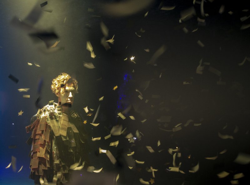 Performer with reflective, angular costume in a dark, smoky space with falling confetti, lit by a spotlight.