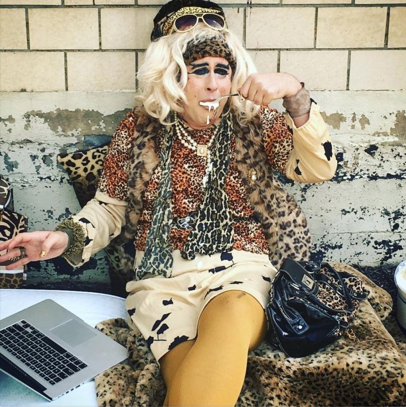 Drag queen in leopard print outfit, blonde wig, and sunglasses, eating with a spoon, laptop nearby.