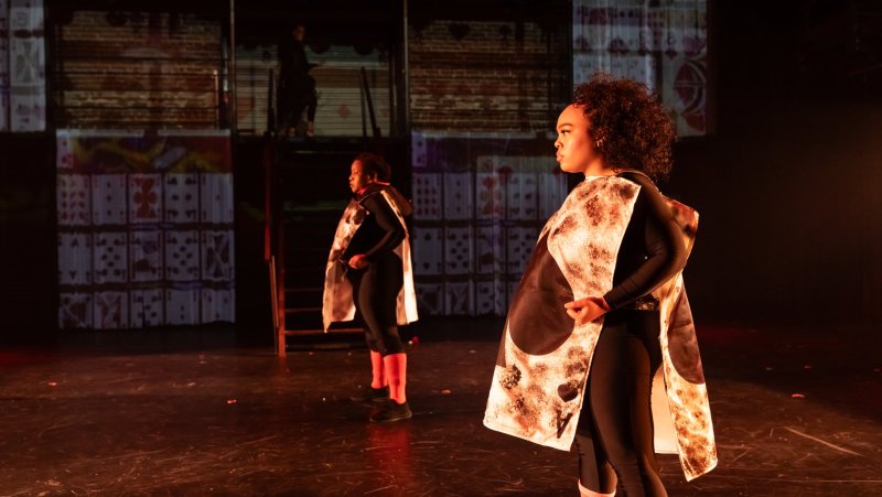 Two figures in black bodysuits and printed capes stand on a stage with playing card projections.