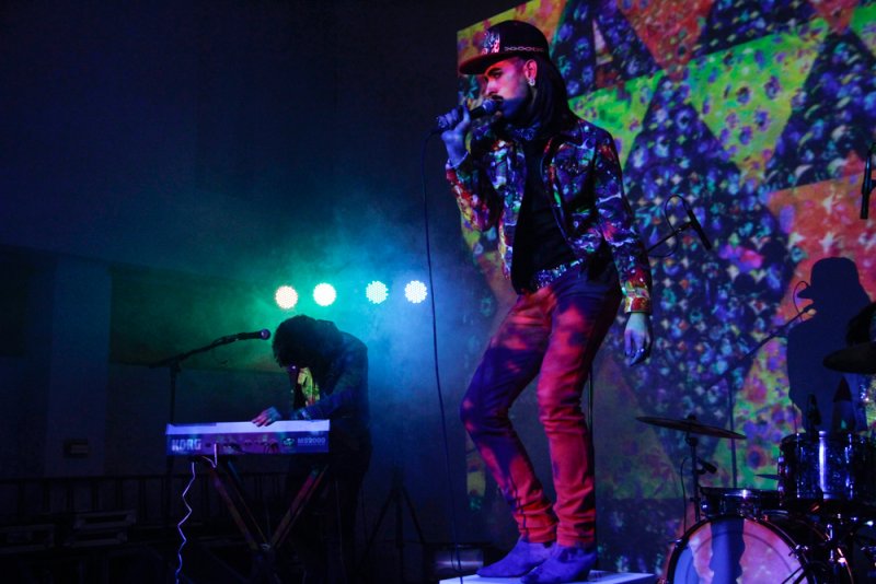 Performance art with musician singing on stage in colorful jacket, keyboardist, and projected backdrop.