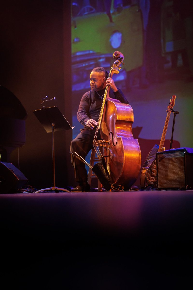 Man playing upright bass on stage with music stand, guitar, and amp; abstract background with car image.