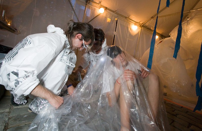 Three people in a makeshift tent, one kneeling, two sitting under clear plastic, illuminated by warm lights.