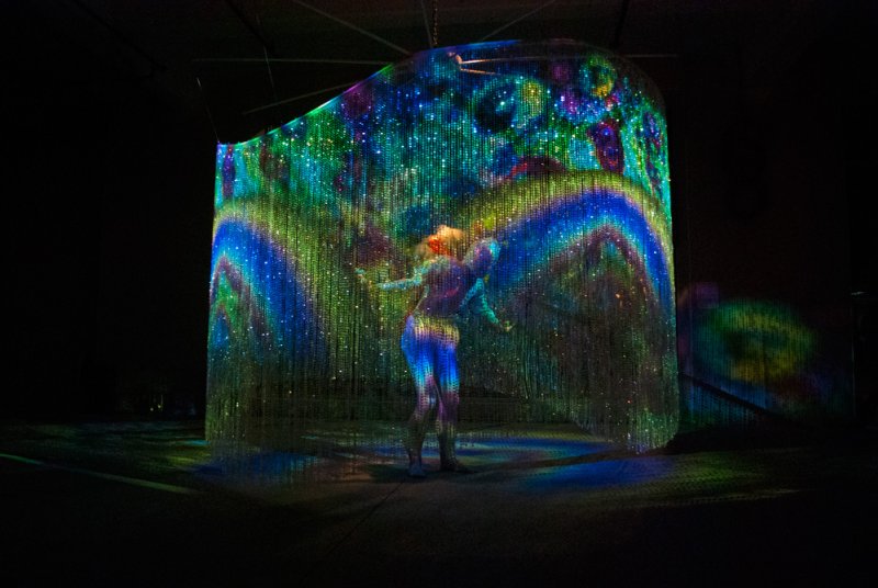 A person stands inside a curved, translucent screen with vibrant, sparkling blue, green, and purple light patterns.