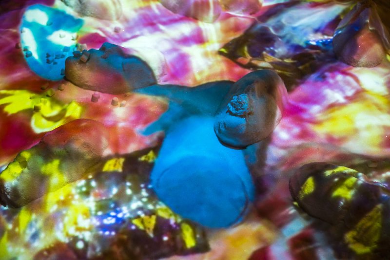 Abstract artwork with textured, rock-like forms in blues, reds, and browns. A vibrant, multicolored light projection illuminates the forms with yellows, pinks, and purples.