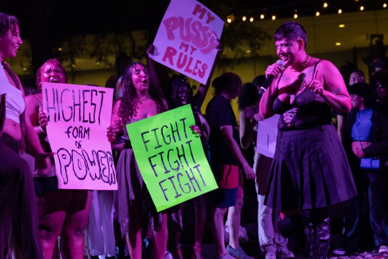 A group of people on a stage with purple and green lighting, holding protest signs. A person in a black pleated skirt and top sings into a microphone.