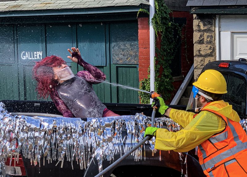 A person in drag with red hair and a face shield is sprayed with water by a person in a yellow hard hat and safety vest.