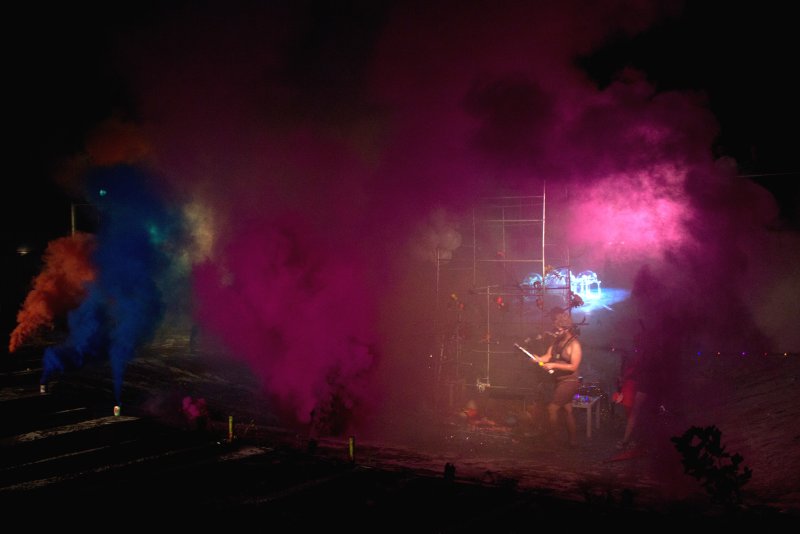 Dark scene with a person in a headdress playing a stringed instrument, surrounded by vibrant pink, blue, and orange smoke.