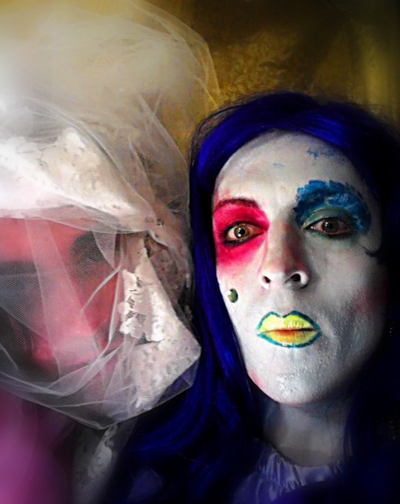 Close-up portrait of a person with white face paint, blue hair, and brightly colored makeup around the eyes and lips. A sheer, lacy veil partially obscures another figure in the background.