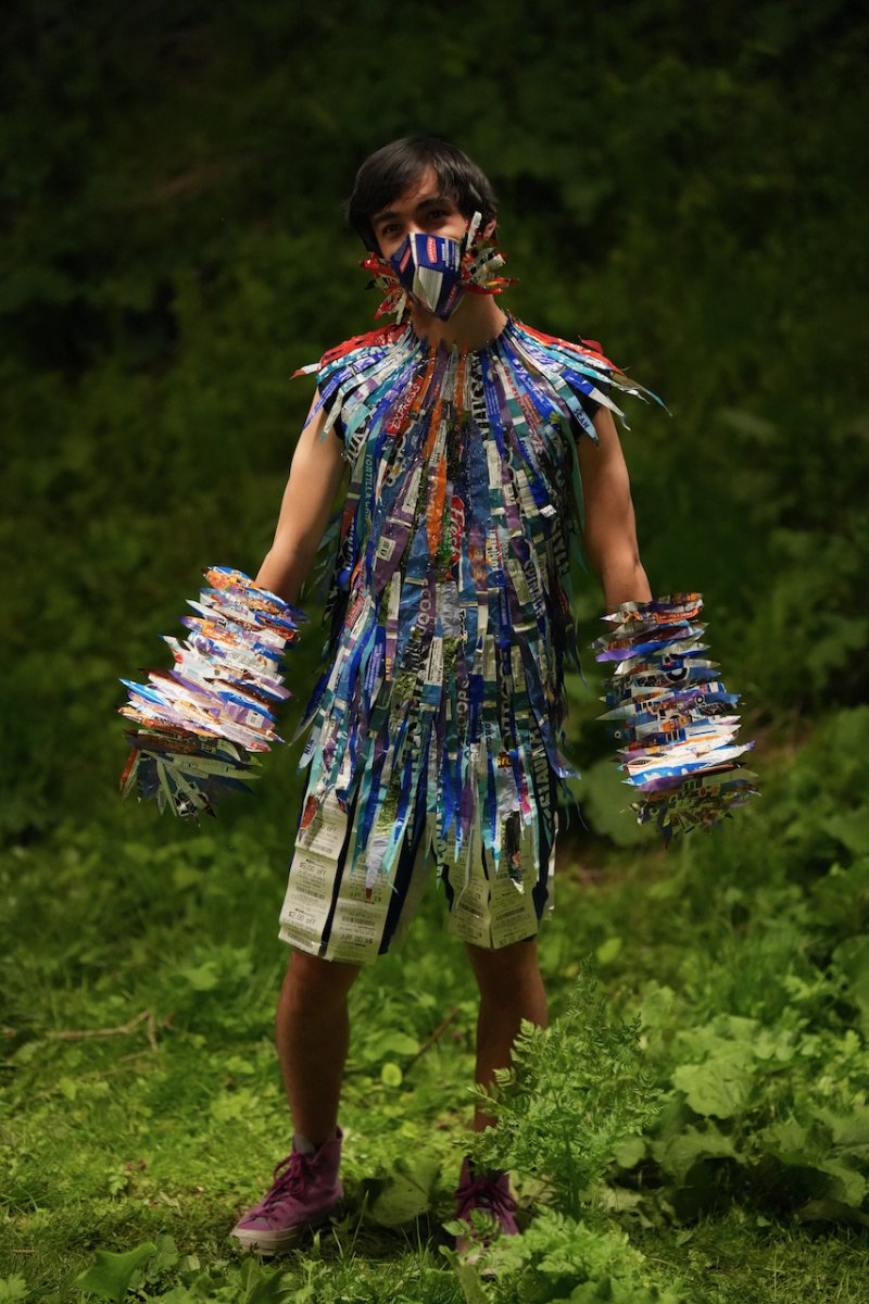 Man in a colorful costume made of shredded paper and a matching mask, standing in a lush green forest.