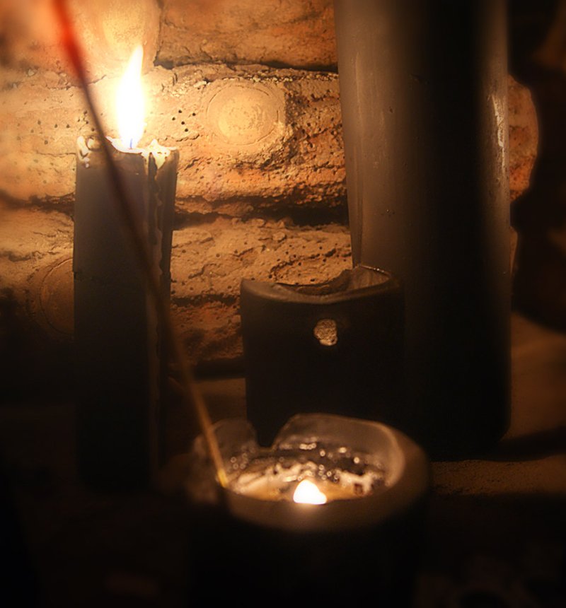 Dark, close-up shot of a lit candle and a small dish with a flame, casting warm light on a brick wall.