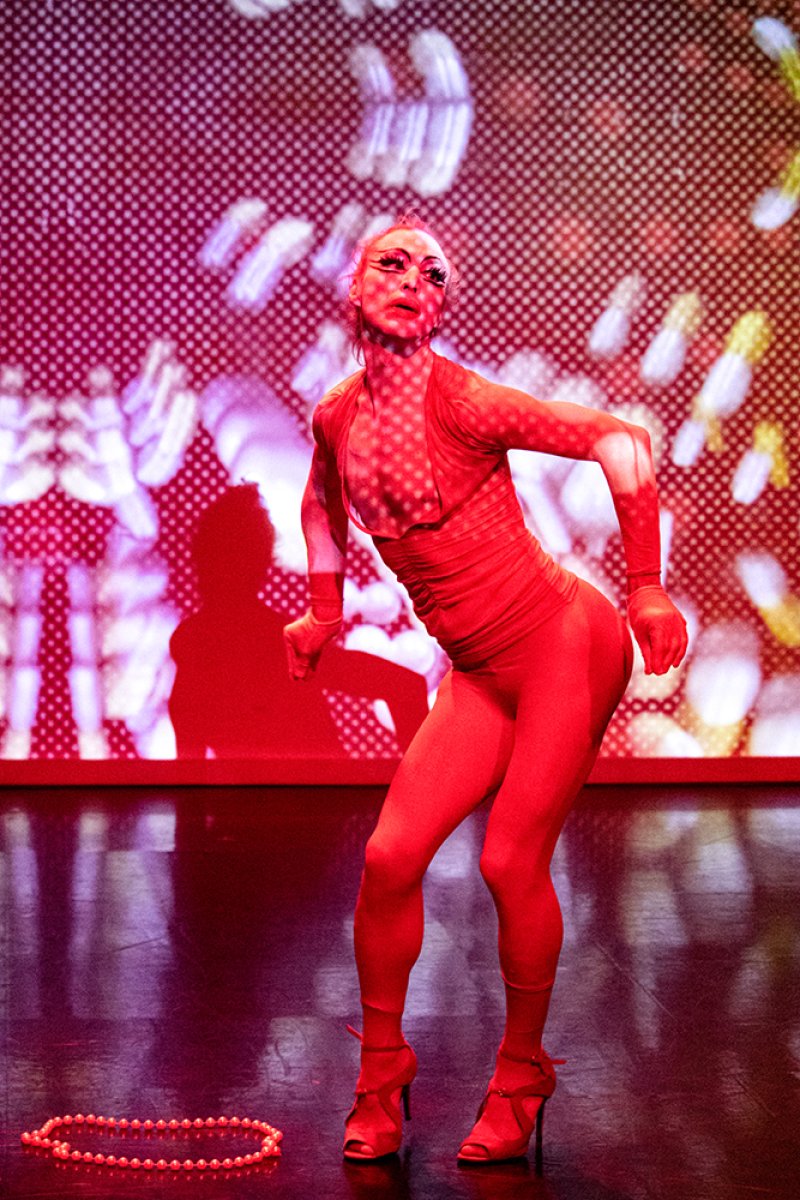 Performer in a red bodysuit and heels striking a pose on a stage with a red and white patterned background.
