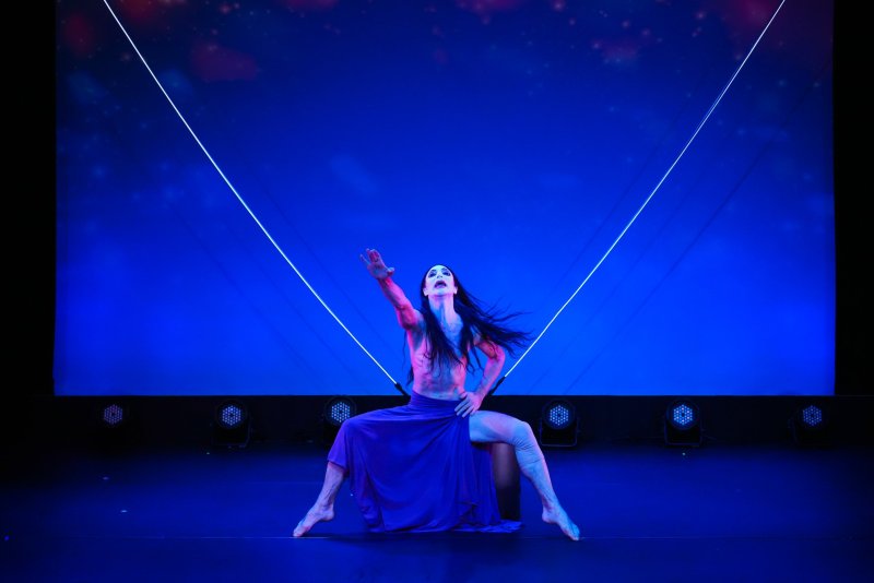 Performance art: dancer in purple skirt poses on stage with blue lighting and geometric laser backdrop.