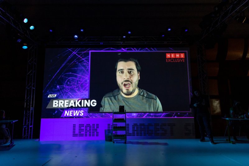 Large screen displays a "Breaking News" segment with a man's face. The screen is framed with purple geometric patterns and the words "News Exclusive." The stage is lit with blue and purple hues.