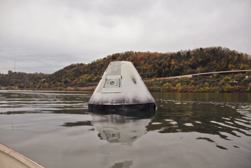 White conical capsule with a small square window floating on calm water, with a tree-covered hill in the background.