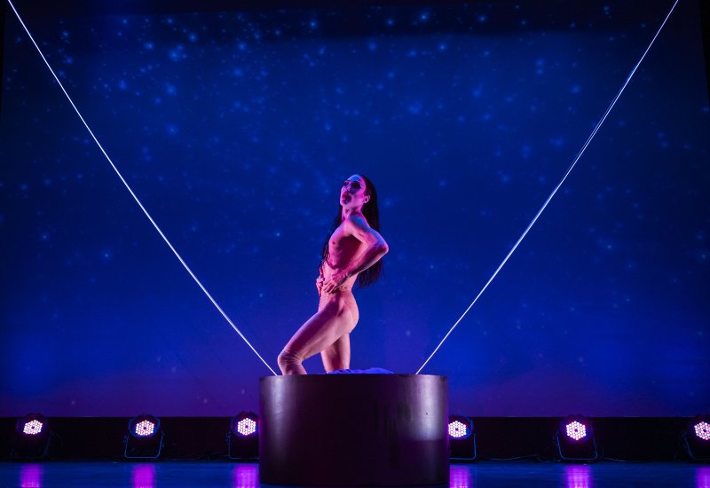 Performer in nude bodysuit stands in a cylinder against a blue starry backdrop with laser lines.