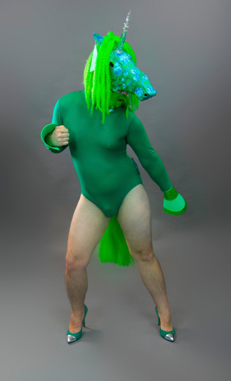 Person in a green unicorn head, green bodysuit, and green heels, posing against a gray background.