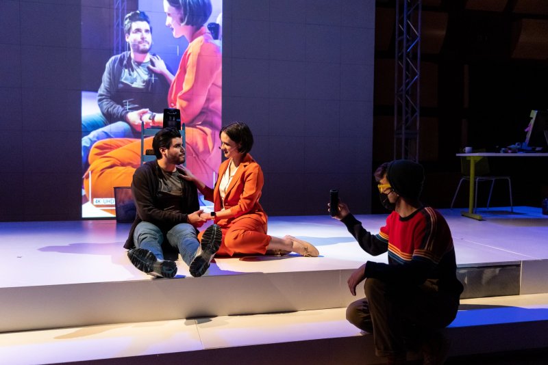 Stage scene with two actors sitting on a white platform, a large screen behind them showing a video of a couple. A third actor kneels to the side, filming with a phone. Predominant colors are orange, blue, and white.