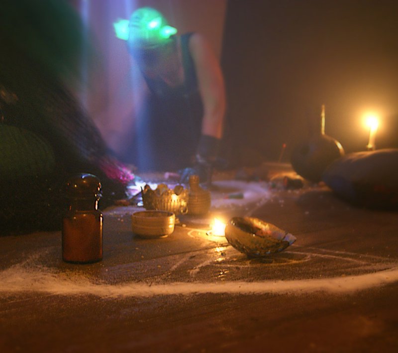 A dark, low-angle shot of a ritualistic scene with a white pentagram drawn in powder, candles, and bottles.