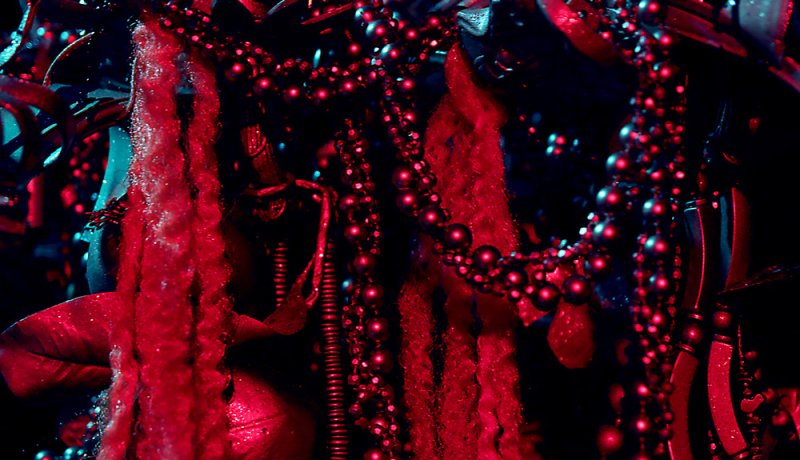 Close-up of a sculpture with red beaded strands, textured red fabric, and a dark background with blue highlights.