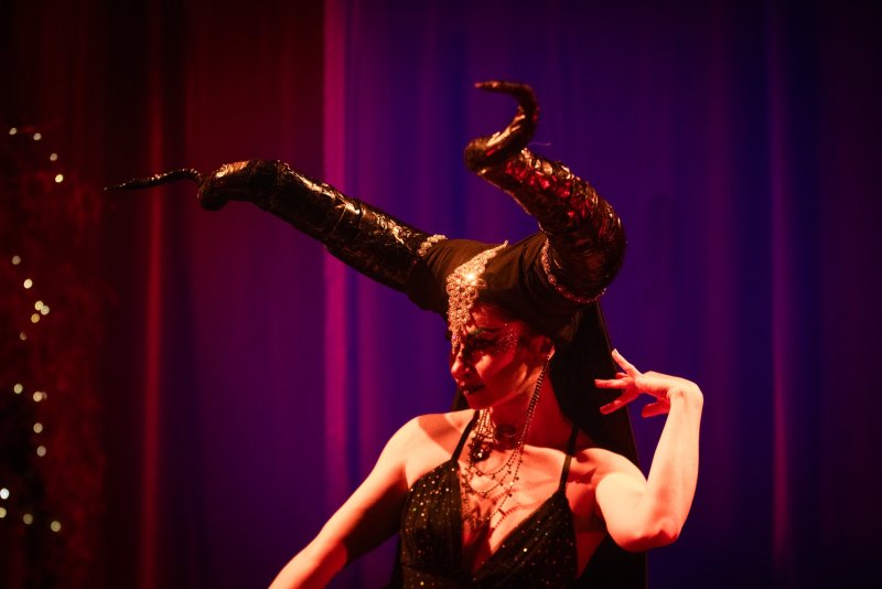 Performer in a black sequined dress and elaborate horned headdress with glitter makeup, lit by red and purple stage lights.