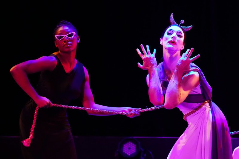 Two figures in theatrical costumes, lit with pink and purple, hold a rope against a black background.