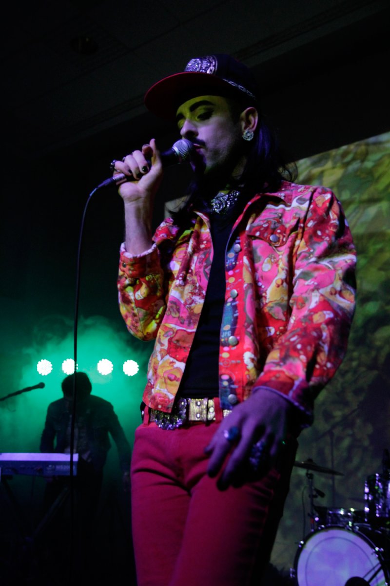 Performance shot: Singer in red pants and patterned jacket sings into a microphone on a stage with green lighting.