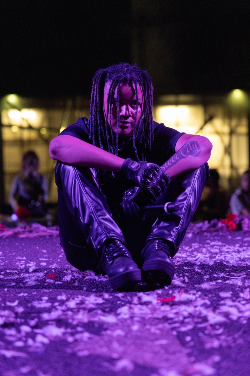 Performer with dark dreadlocks, black shirt, and shiny black pants sits on a floor covered in purple and white confetti, illuminated by purple stage lights.