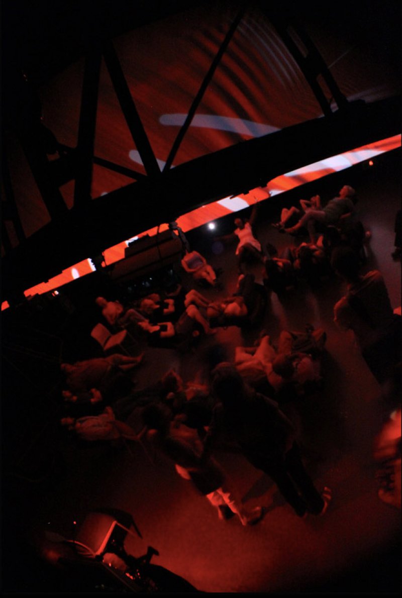 Dark, high-angle shot of people lying on a red-lit floor, with abstract red and white projections on a slanted ceiling.