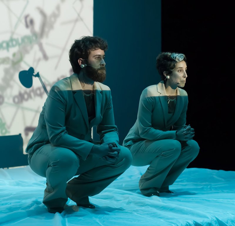 Two performers in light blue suits crouch on a white sheet, with text projected behind them.