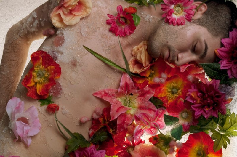 Man covered in flowers and glitter, eyes closed, surrounded by vibrant red, pink, and orange blossoms.