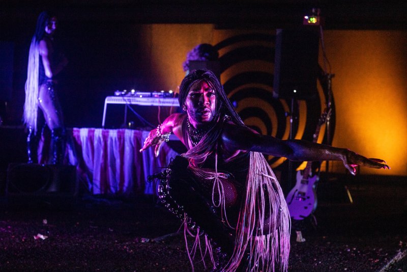 Performance shot with a central figure in a dramatic pose, illuminated by purple and orange light. Braids, jewelry, and stage equipment are visible, with a figure in the background.